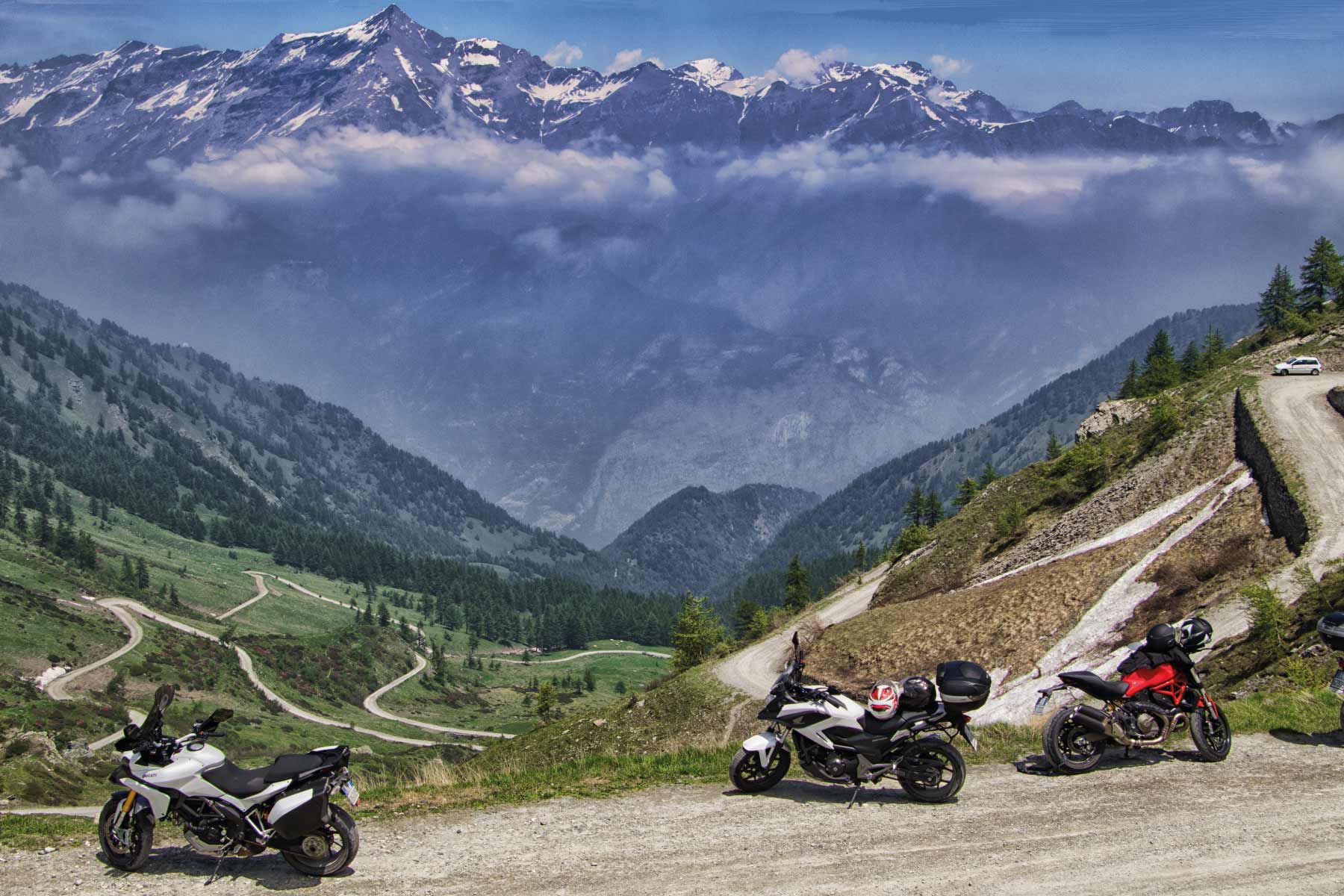 Motociclette parcheggiate con vista su strade serpeggianti e montagne nebbiose.