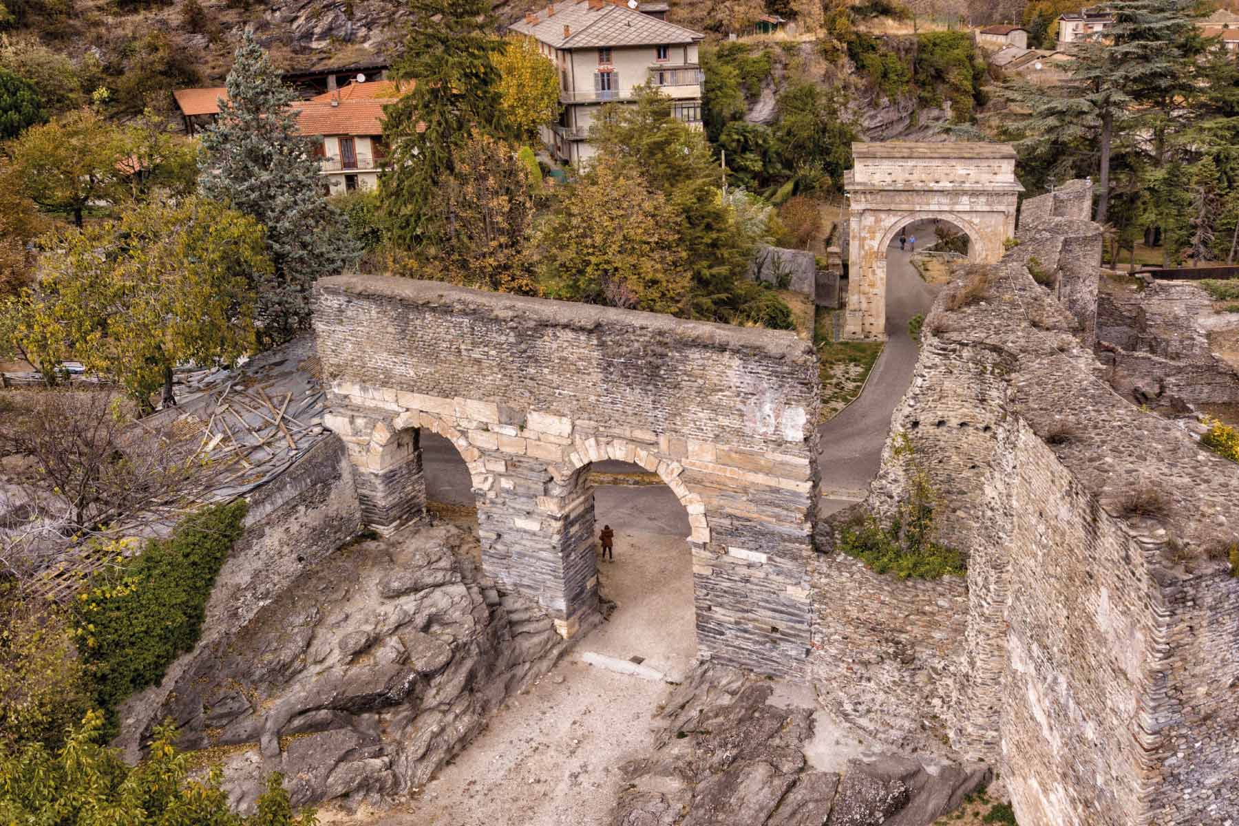 Antico arco di pietra e rovine in una zona residenziale con vegetazione.