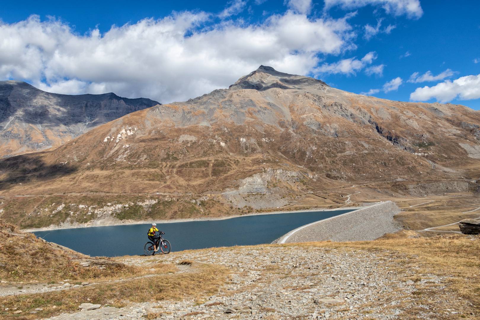 Ciclista su strada di montagna vicino a lago turchese e innevate vette.