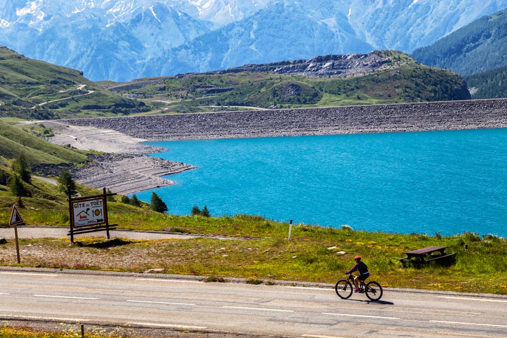 Ciclista su strada di montagna vicino a lago turchese e innevate vette.