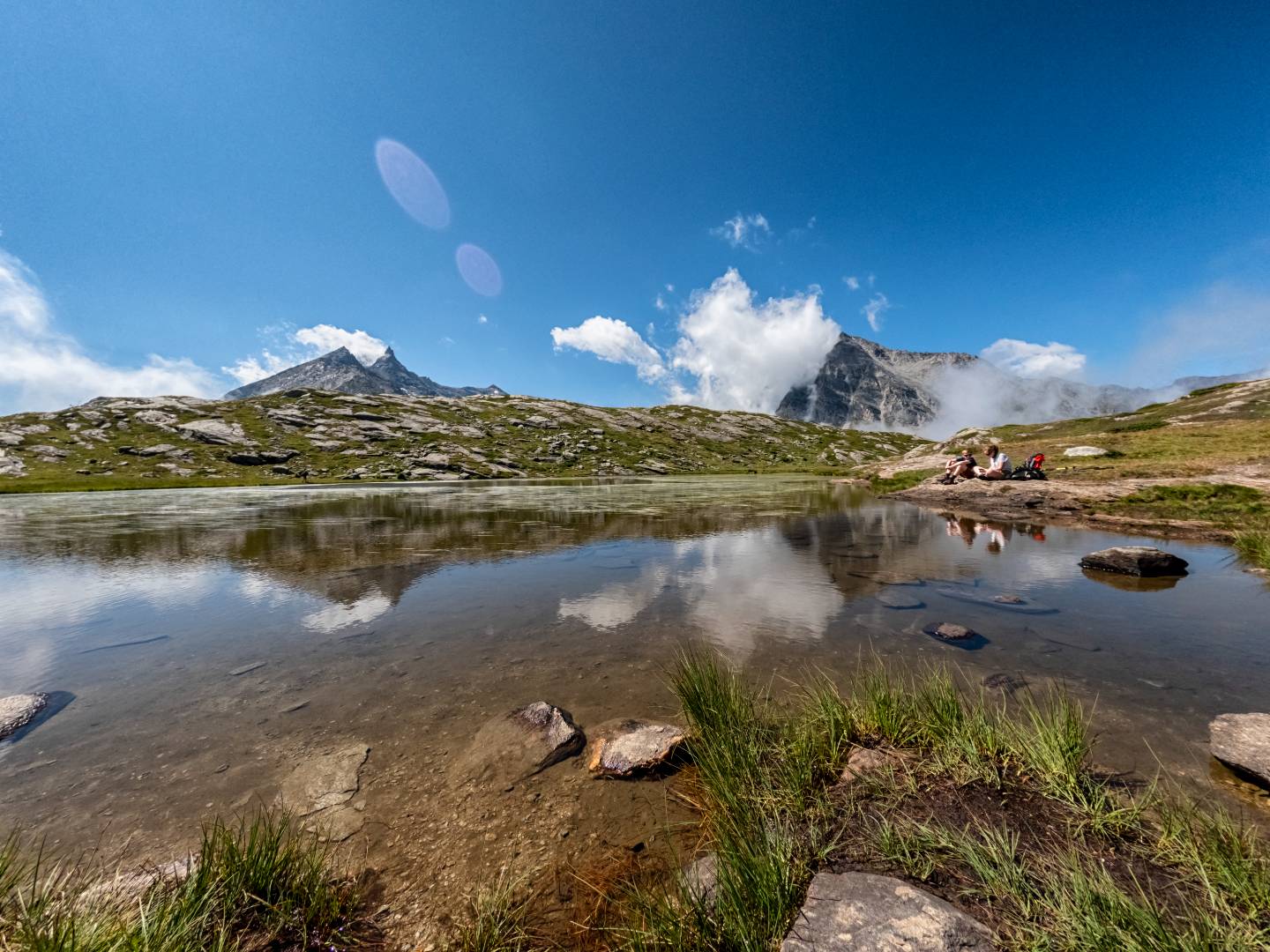 Laghetto alpino circondato da erba e montagne rocciose sotto cielo azzurro.