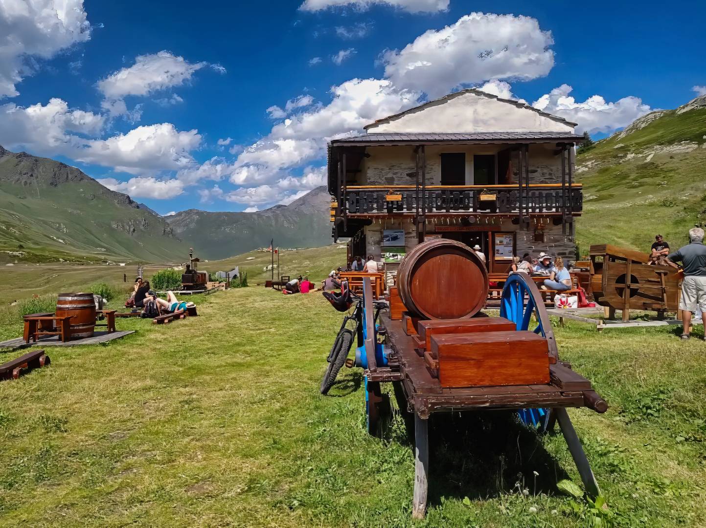 Area picnic in montagna con persone, bici, casetta in legno e montagne.