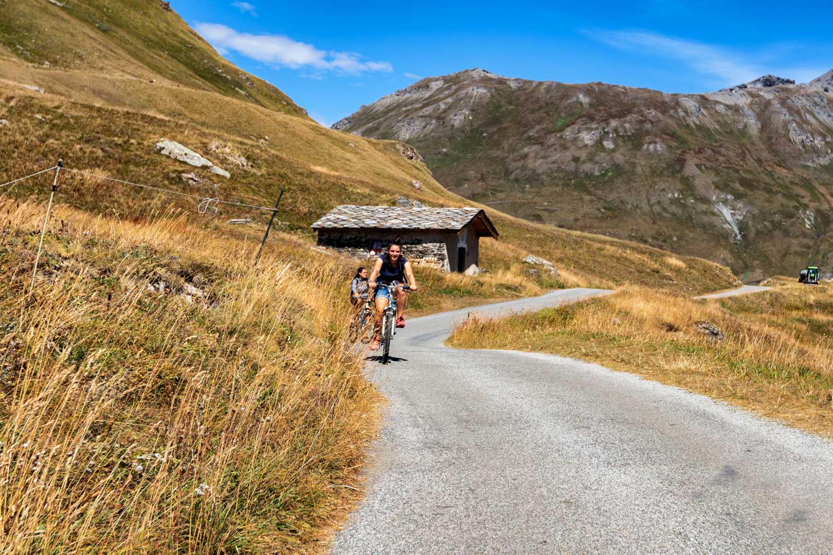 Ciclisti su strada di montagna con erba secca e piccola baita.