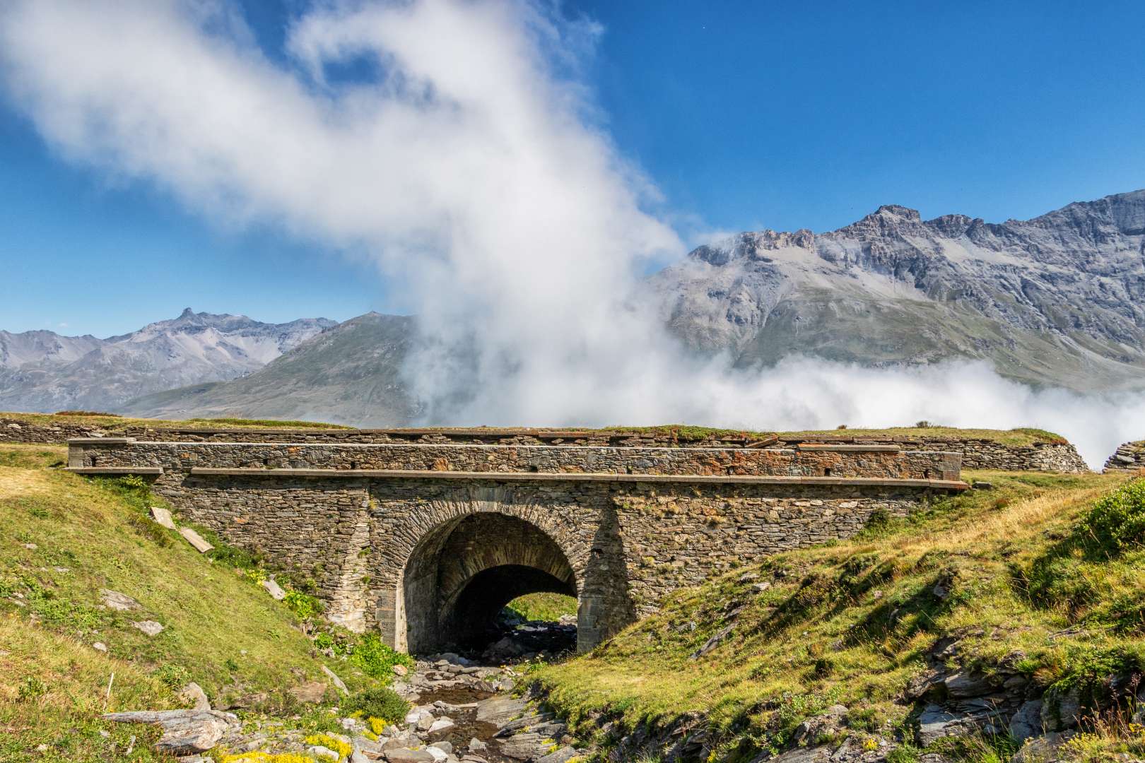 Ponte in pietra in montagna, nuvole basse e vette in lontananza.