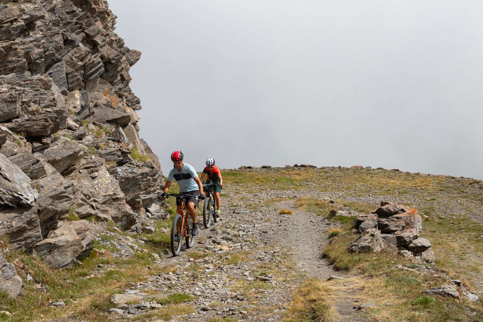 Ciclisti in mountain bike su sentiero alpino con parete rocciosa e cielo nebbioso.