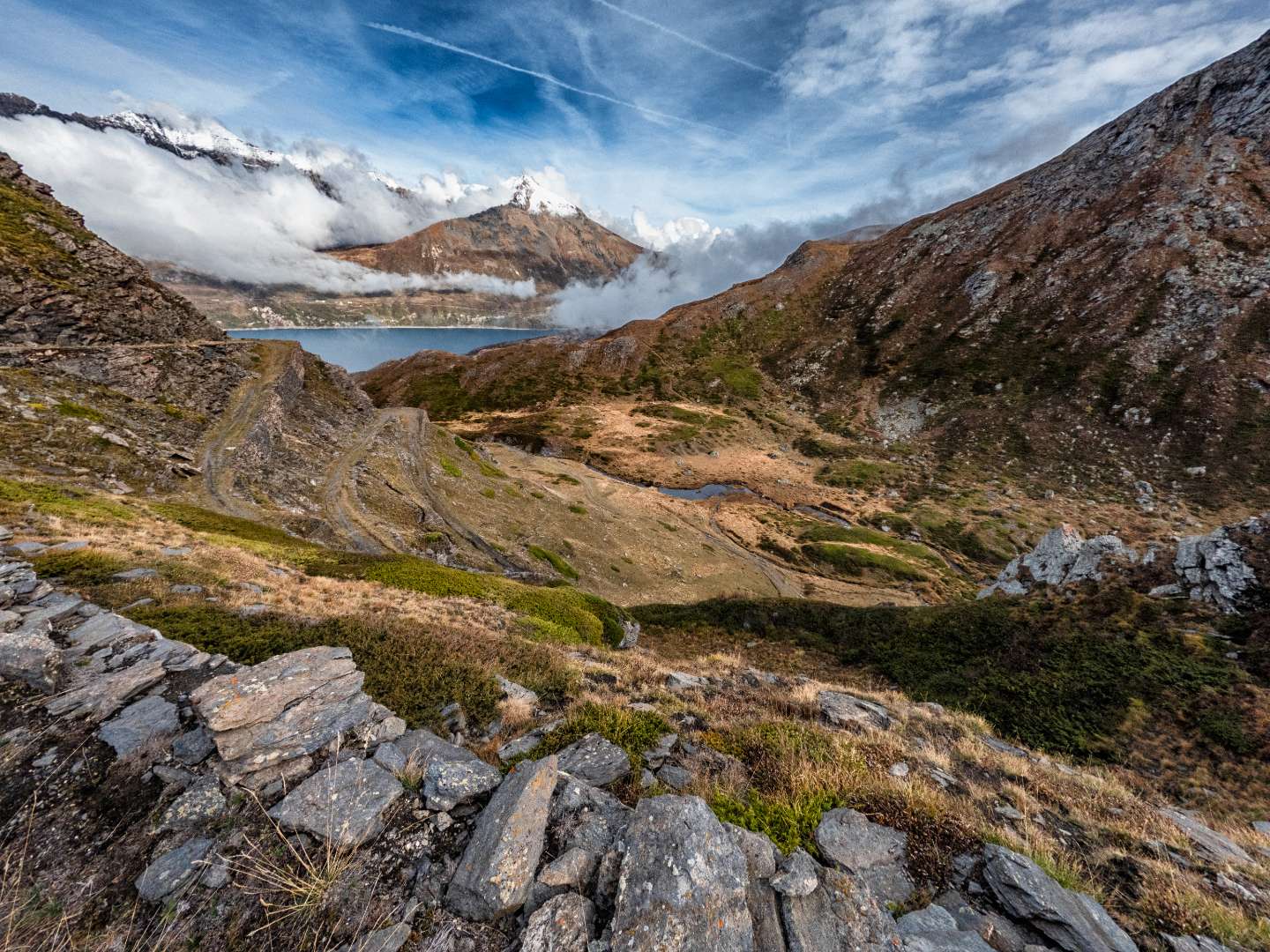 Percorso montano roccioso che si snoda verso un lago circondato da nuvole.