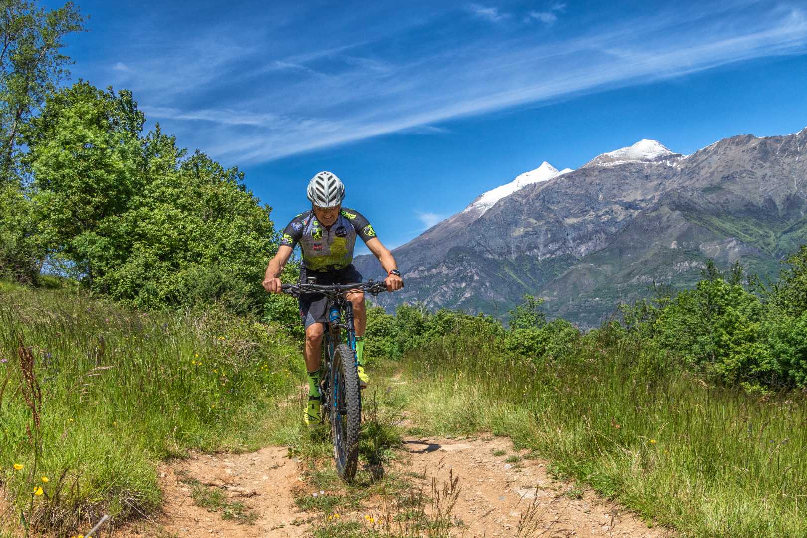 Biker in azione su sentiero di montagna con cime innevate sullo sfondo.