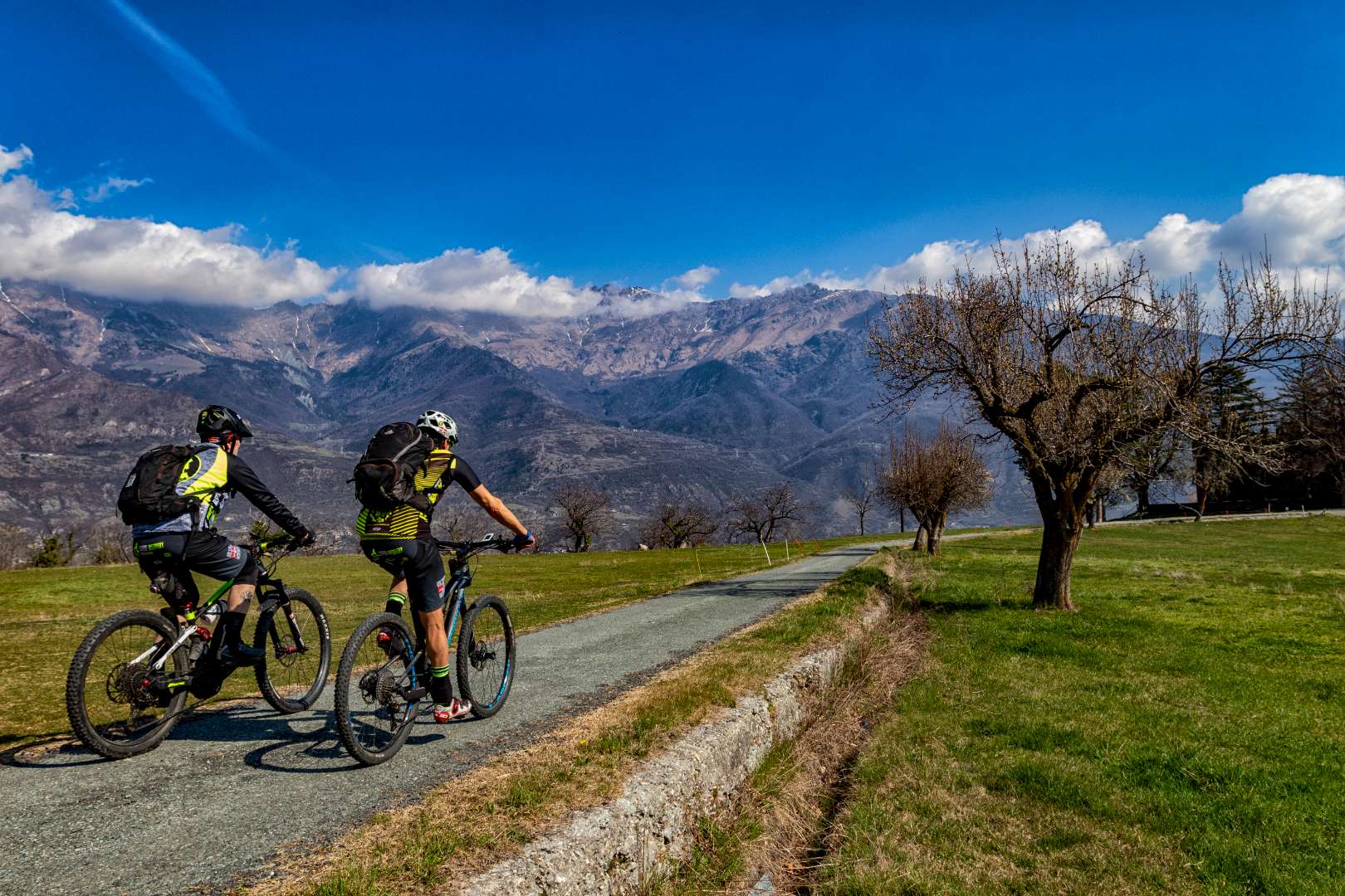 Ciclisti in mountain bike su percorso erbose con vista montagne e cielo azzurro.