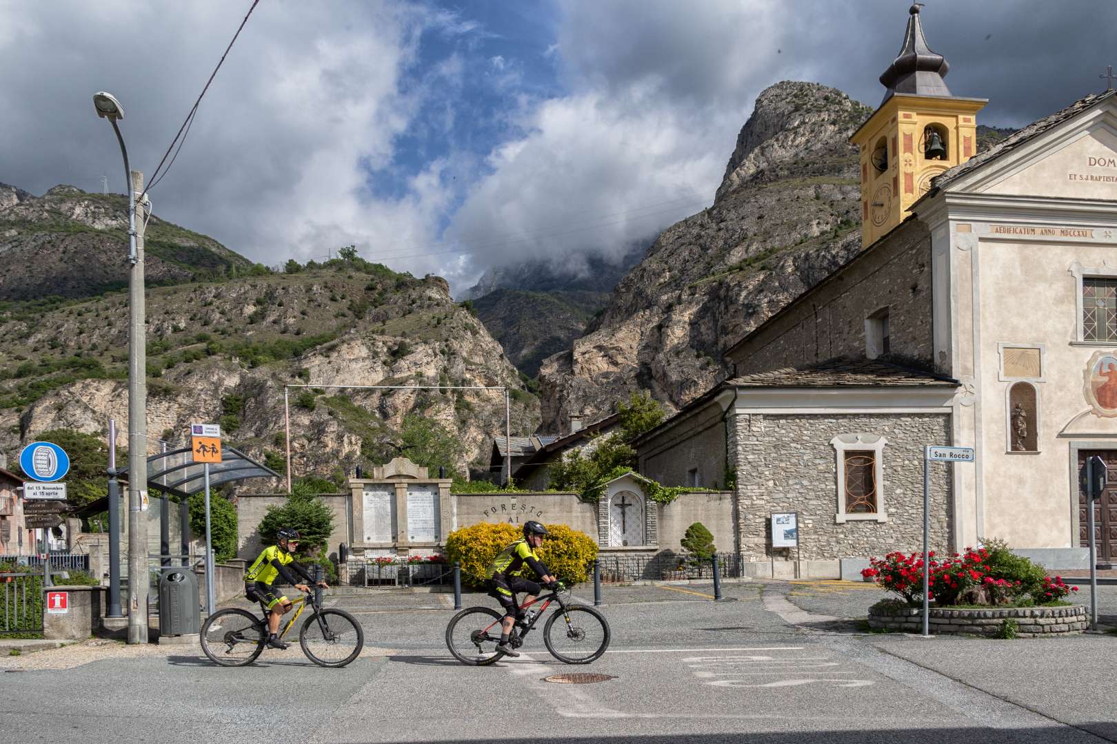 Ciclisti passano davanti a chiesa in paesino montano sotto cielo nuvoloso.