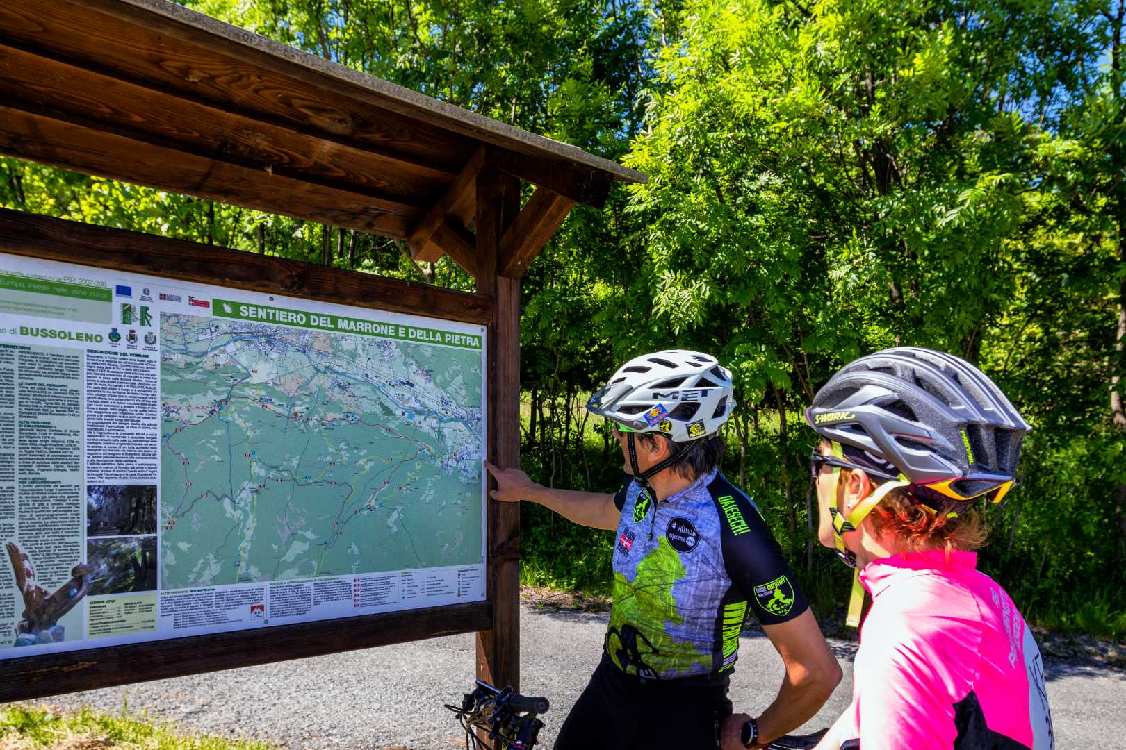 Ciclisti consultano cartello informativo di sentiero escursionistico in ambiente boschivo.